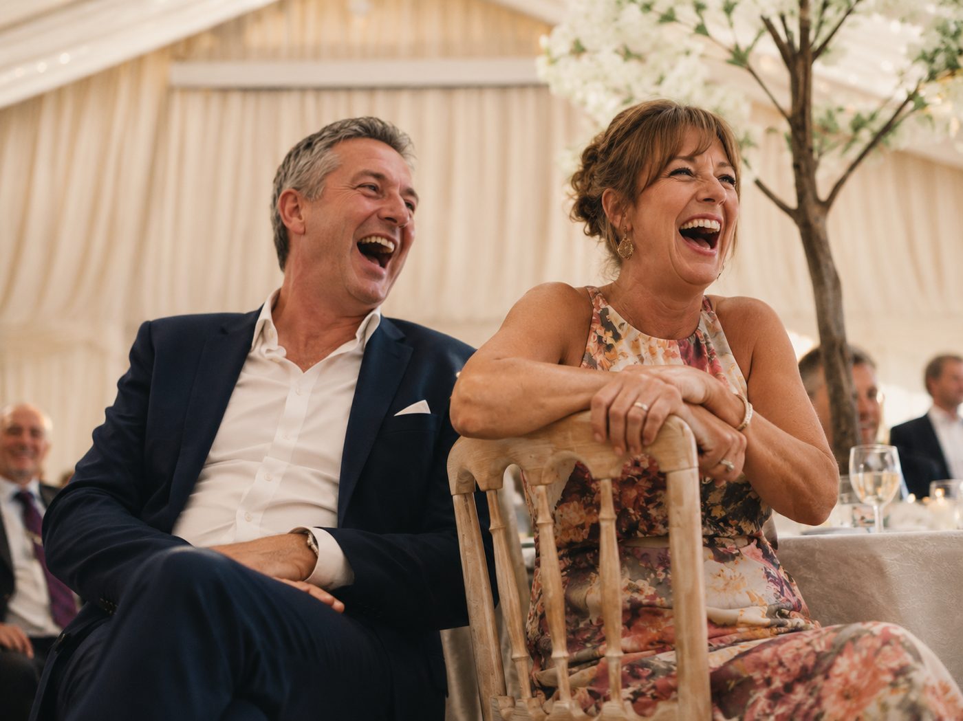 Two wedding guests laughing hard together at a reception table under a marquee