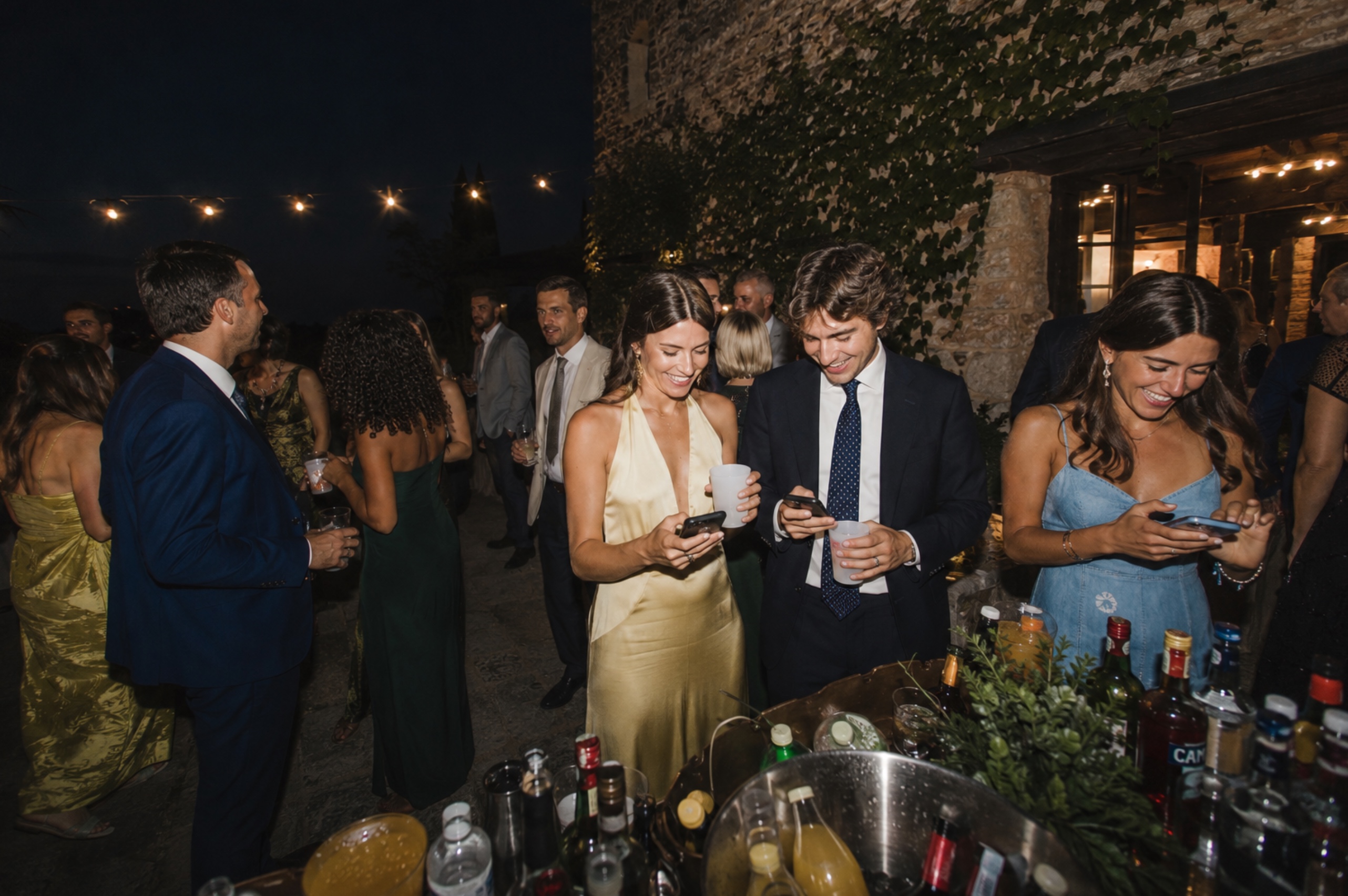 Wedding guests laughing and looking at phones during a nighttime outdoor cocktail reception