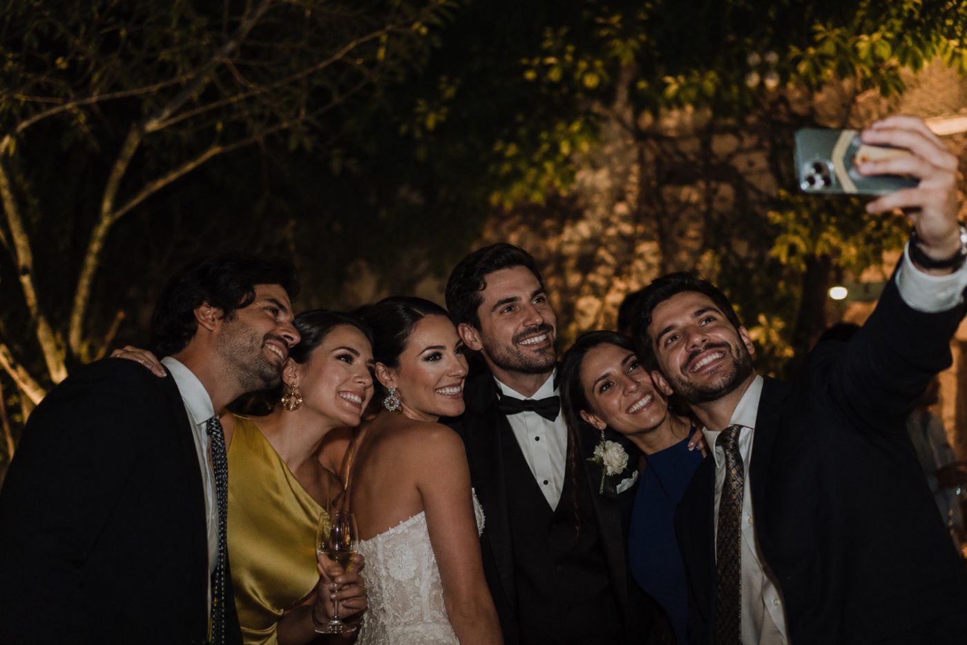 Bride and wedding party squeezing in for a group selfie on phones at a nighttime reception