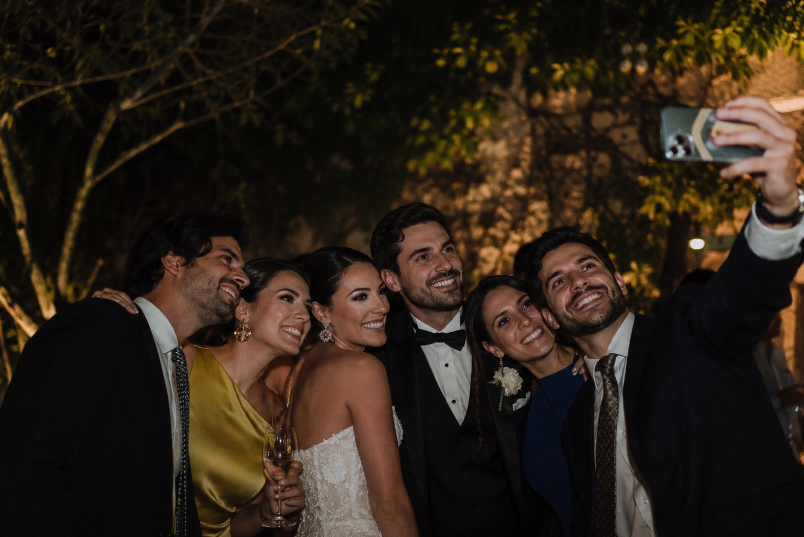 Wedding party huddled for a night-time group selfie, smiling at a phone