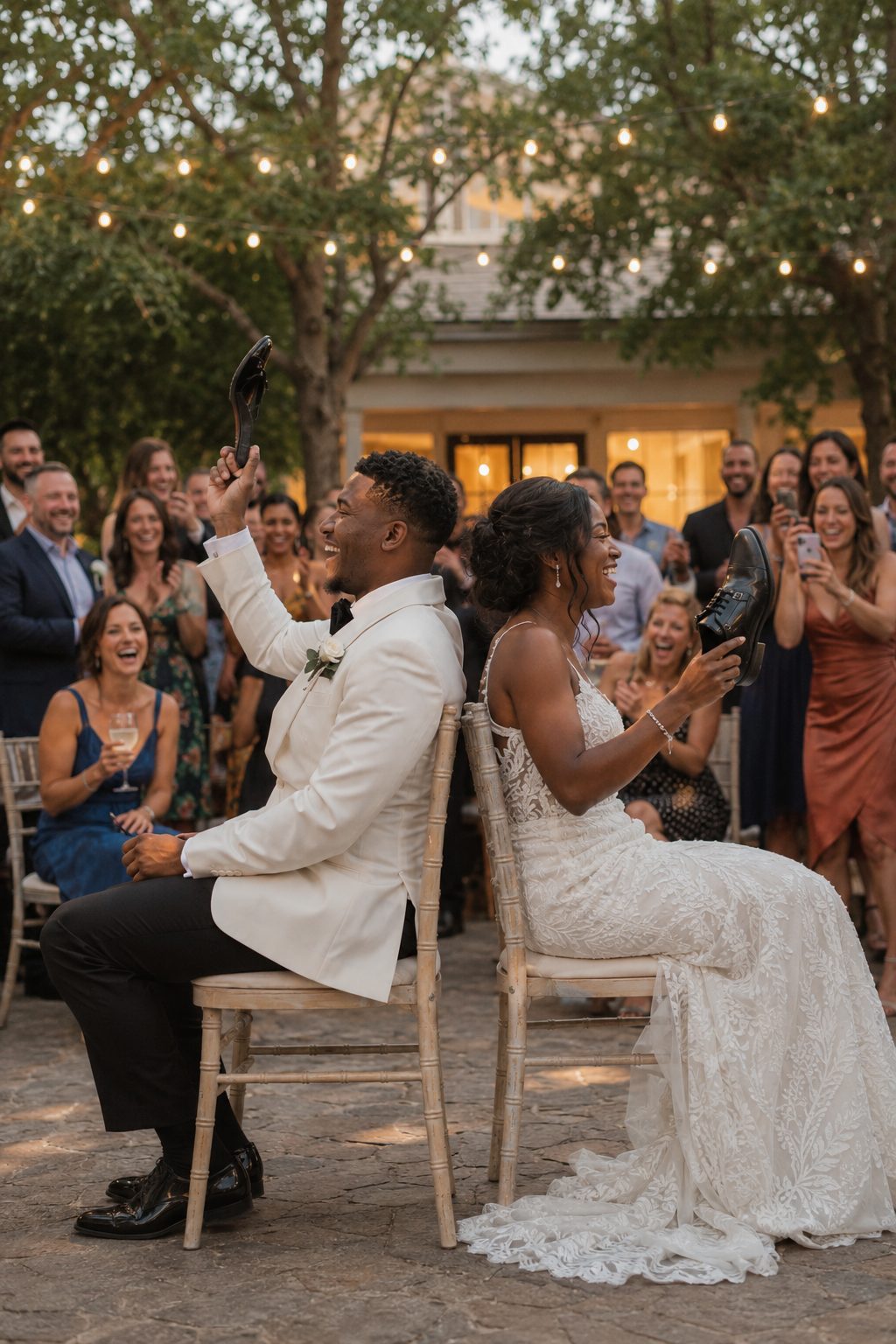 Bride and groom sitting back-to-back laughing while holding up shoes during the wedding shoe game